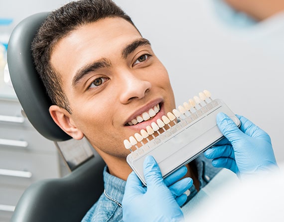 a young man sits in a chair while a dental technician compares his teeth to a whitening chart